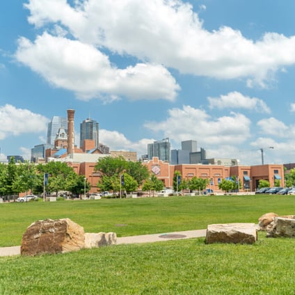 Denver skyline viewed from a grassy area, with large rocks in the foreground, trees and the Tivoli building in the middle ground, and downtown high-rise buildings beneath a bright blue sky with clouds.