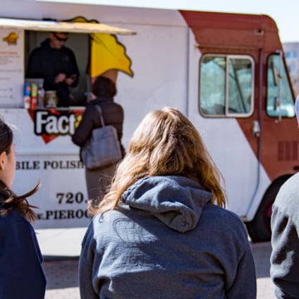 Three people sit with their backs to the camera facing a food truck serving Polish food. Menus and food photos are displayed on the truck, and another truck is parked nearby.