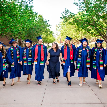Nine graduates in blue commencement regalia walk across a campus pathway lined with green trees. Many wear colorful stoles and graduation cords. A person in a navy dress walks in the middle of the group as they move toward the camera.