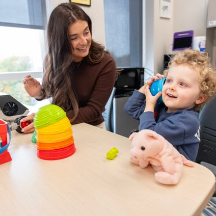 A smiling woman sits beside a child at a table in a therapy room. The child holds a blue cup-shaped object to their ear while looking toward the camera, with a pink stuffed animal on the table. Colorful stacking toys are arranged nearby.