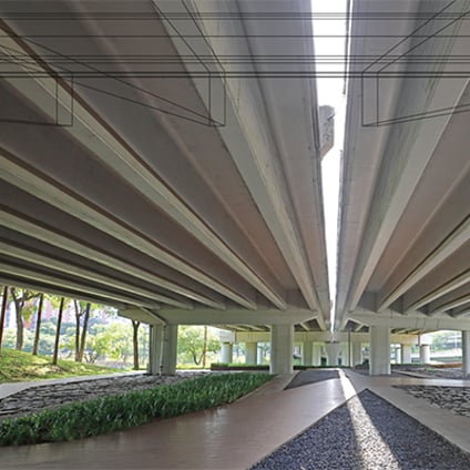 A landscaped pedestrian walkway runs beneath two parallel elevated concrete roadways, with structural columns lining both sides. Long bands of greenery, stone landscaping, and trees border the walkway.