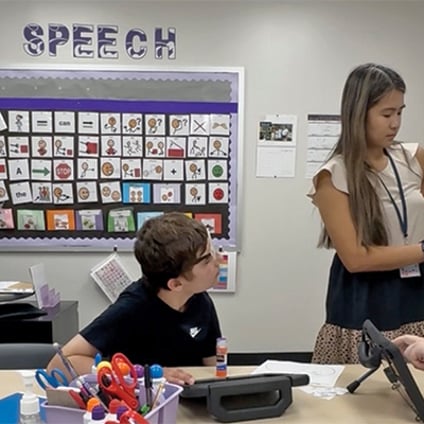 A speech therapy session in a classroom. A child sits at a table looking toward a woman who holds a tablet displaying an activity. A speech-sound picture board labeled “SPEECH” covers the wall behind them.
