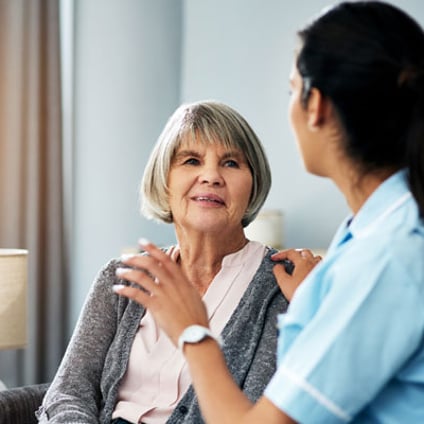 AAn older woman sits facing a female health care professional in a blue uniform who is speaking with her and resting a hand on her shoulder. They are seated in a bright, home-like room with a lamp and curtains in the background.