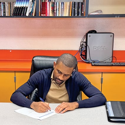 SLP Sybrey “Sunny” Callwood sits at a desk in a classroom, writing on a worksheet with a pen. He wears a dark cardigan over a tan shirt, with a black binder beside him. Behind him are orange and yellow cabinets, a desktop computer, and shelves filled with books.
