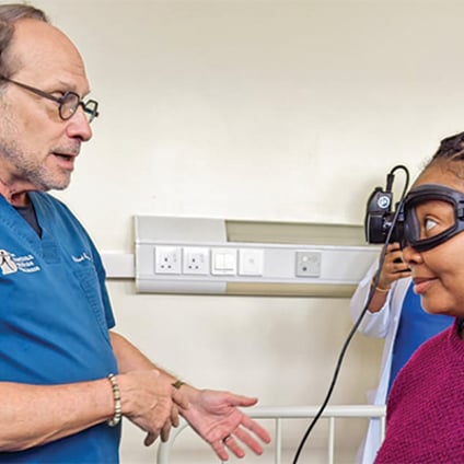 A health care professional in blue scrubs speaks with a woman wearing protective goggles connected to a medical device. They stand in a clinical examination room with medical equipment and electrical outlets visible on the wall behind them.