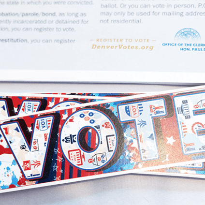 A stack of colorful “VOTE” postcards sits on a voting information sheet from the Denver Clerk and Recorder’s Office. The sheet includes the voter registration website DenverVotes.org, while the postcards feature red, white, and blue graphics.