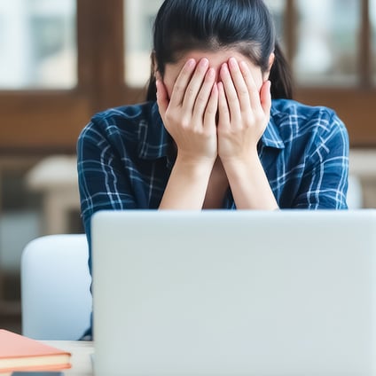 A person sits behind a laptop at a desk, covering their face with both hands. Pencils, notebooks, and a small plant are on the desk, with books in the background.