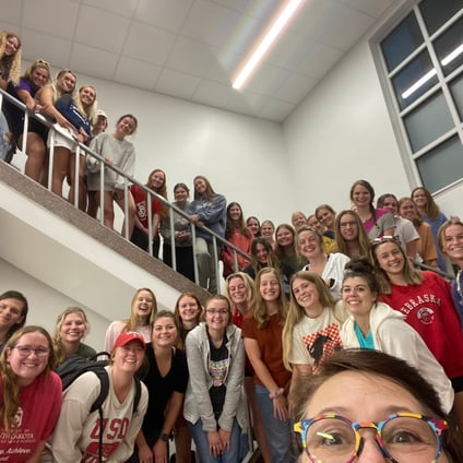 A large group of smiling students poses on and around a stairwell inside a school building, with some standing on the upper landing and others gathered on the stairs and lower level. A person wearing colorful glasses takes the selfie from the foreground. Several students wear college-branded clothing and backpacks.